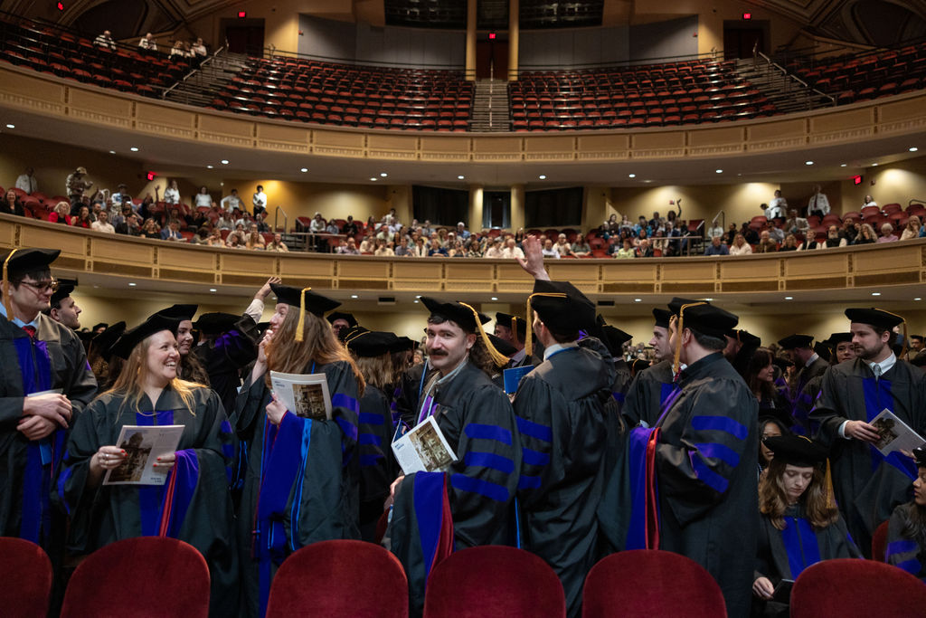 Photo of graduates in robes and tams in the auditorium standing, smiling, and chatting.