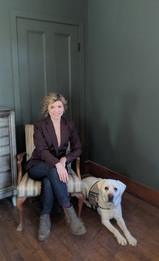 Photo of woman seated in chair with service dog lab lying next to her. Both are looking at the camera.