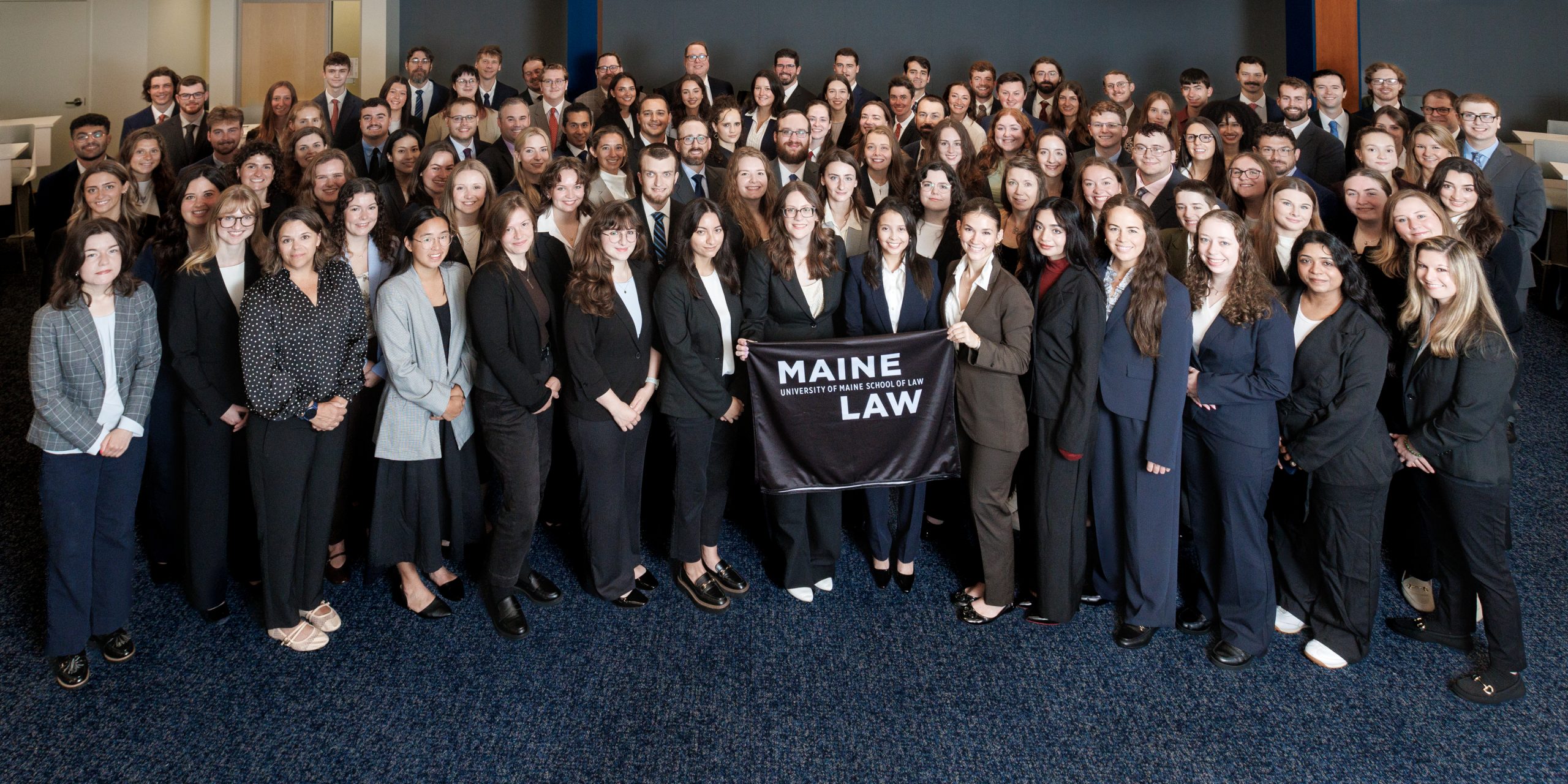 A photo of the Class of 2029 taken in the Moot Courtroom, students out front hold a Maine Law flag.