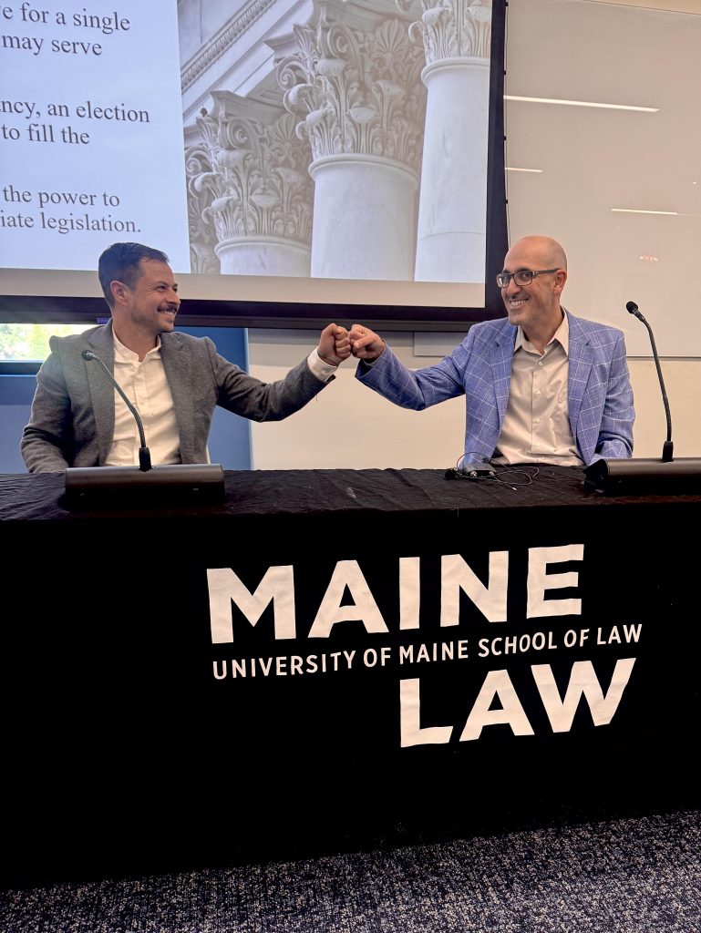 Bloomberg (left) and Bam end their discussion with a friendly fist bump, seated at the front of a classroom with their power point in the background.